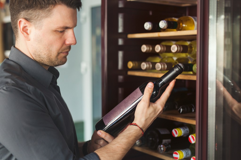 il-sommelier-sceglie-una-bottiglia-di-vino-vintage-invecchiato-vicino-all-armadio-frigorifero-aperto-in-un-negozio-di-vini-contemporaneo